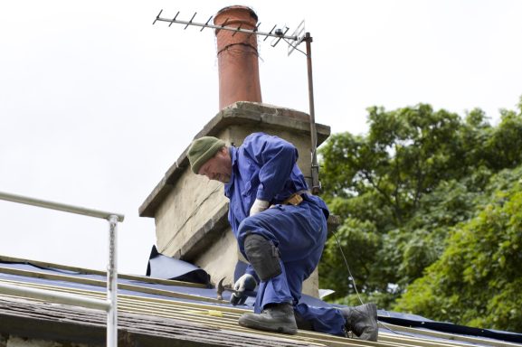 Handwerker in blauer Arbeitskleidung auf dem Dach, in der Nähe eines Schornsteins.