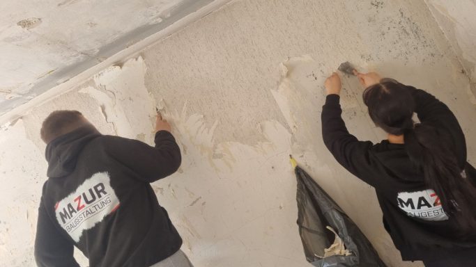 Zwei Mitarbeiter der Firma Mazur Baugestaltung entfernen Tapete von einer Wand in einem renovierungsbedürftigen Raum.