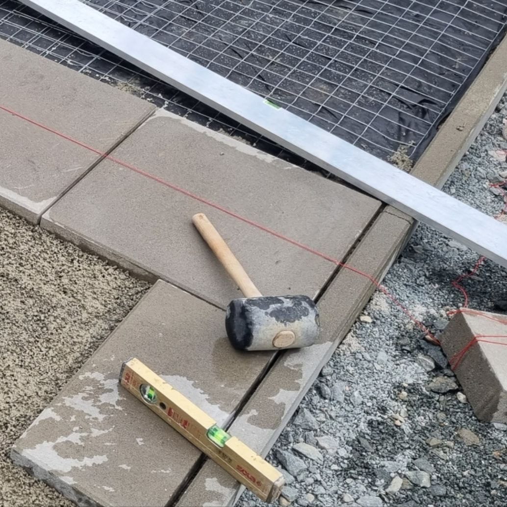 Ein Gummihammer liegt neben verlegten Platten und einem Wasserwaage auf einer Baustelle.