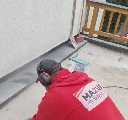 Handwerker in roter Jacke arbeitet auf einem Balkon, trägt Gehörschutz.