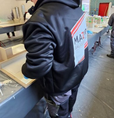 Person steht an einem Tisch und bearbeitet eine Oberfläche, trägt eine schwarze Jacke mit Logo.